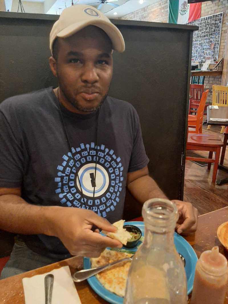 A photo of Mark-Shane Scale in a restaurant, wearing a 1Password t-shirt and cap