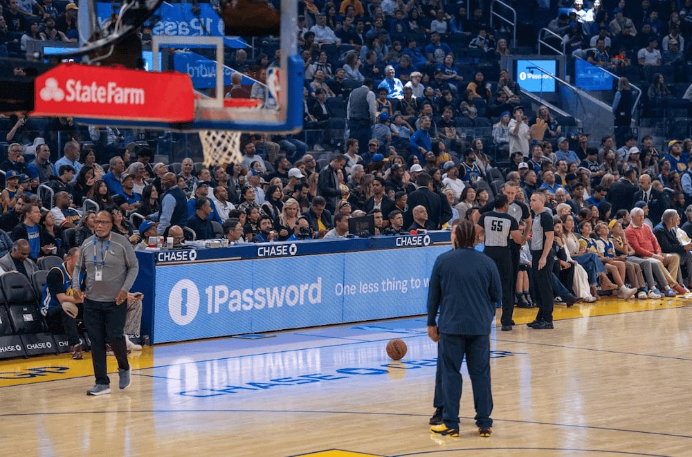 A basketball arena with spectators, a basketball on the court, and an advertisement for 1Password.